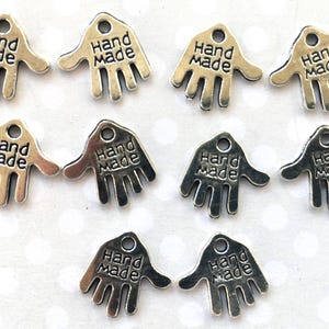 May include: Silver-toned metal charms in the shape of a hand, each with the words "Hand Made" engraved on them. The charms have a small hole at the top for stringing or attaching. Ideal for jewelry making or craft projects.