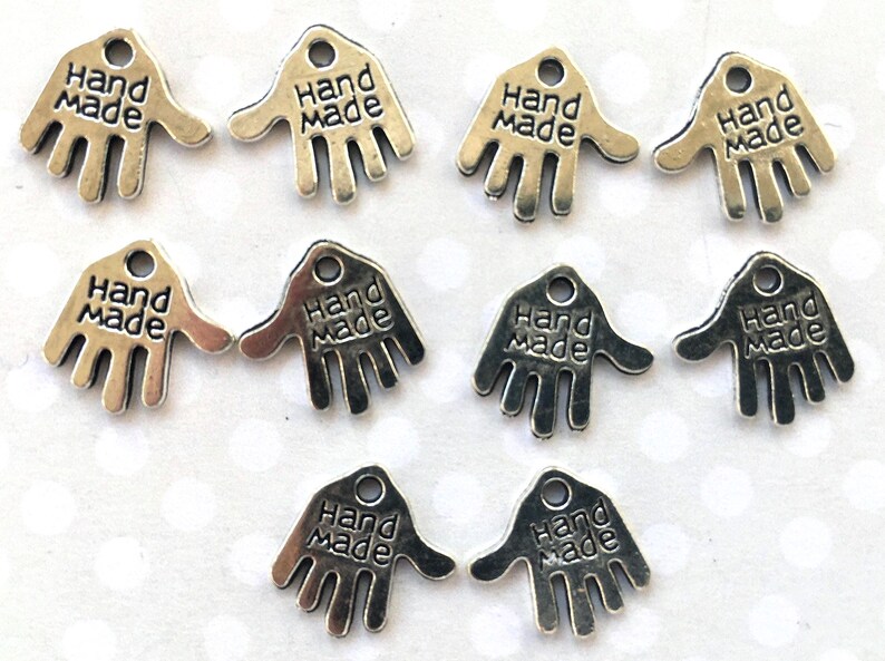 May include: Silver-toned metal charms in the shape of a hand, each with the words "Hand Made" engraved on them. The charms have a small hole at the top for stringing or attaching. Ideal for jewelry making or craft projects.