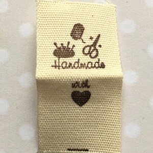 K&ouml;nnte beinhalten: Ein beigefarbenes Stofflabel mit brauner Tinte. Das Label lautet "Handmade with" und hat ein Herzsymbol. Au&szlig;erdem sind Bilder einer Garnrolle, einer Nadel und einer Schere zu sehen.