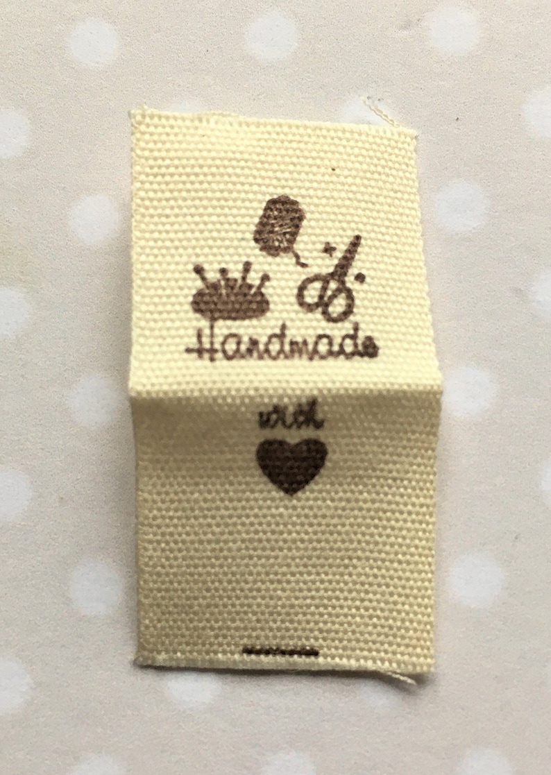 K&ouml;nnte beinhalten: Ein beigefarbenes Stofflabel mit brauner Tinte. Das Label lautet "Handmade with" und hat ein Herzsymbol. Au&szlig;erdem sind Bilder einer Garnrolle, einer Nadel und einer Schere zu sehen.