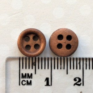 以下が含まれることがあります： 4つの穴が空いた茶色の木製ボタンが2つ。ボタンの直径は約1cmで、定規の隣に置かれています。