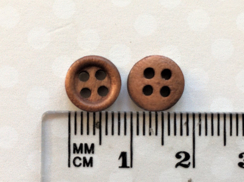 以下が含まれることがあります： 4つの穴が空いた茶色の木製ボタンが2つ。ボタンの直径は約1cmで、定規の隣に置かれています。