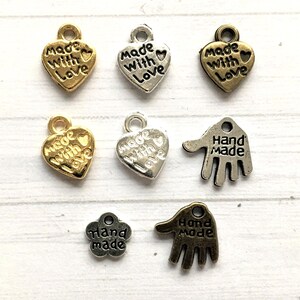 May include: A collection of metal charms in various shapes and finishes. Heart-shaped charms with "Made With Love" text, and hand-shaped charms with "Hand Made" text. Colors include gold, silver, and bronze.