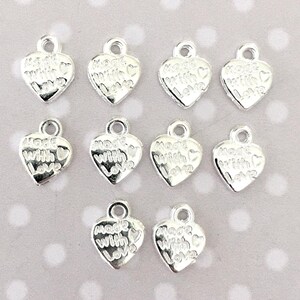 May include: A collection of silver-colored heart-shaped charms. Each charm has a small loop at the top and the words "Made with Love" engraved on the front. The charms are scattered on a light background.