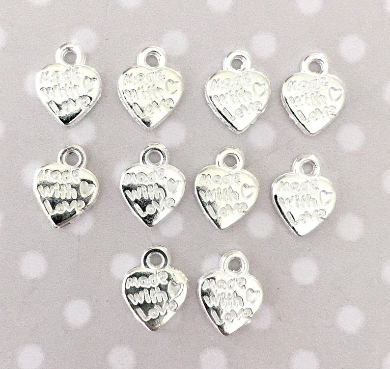 May include: A collection of silver-colored heart-shaped charms. Each charm has a small loop at the top and the words "Made with Love" engraved on the front. The charms are scattered on a light background.