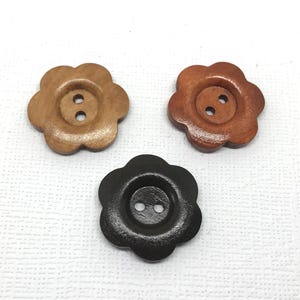 May include: Three flower-shaped wooden buttons in shades of brown and black. Each button features a two-hole design in the centre for sewing. The buttons are displayed on a white textured surface, highlighting their natural wood grain.
