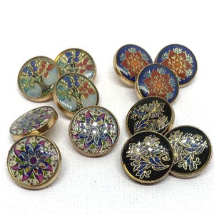 May include: A collection of round metal buttons featuring various colourful enamel designs. The patterns include stylised trees, floral mandalas, and botanical motifs in shades of blue, purple, red, orange, and black with gold-toned metal edges.