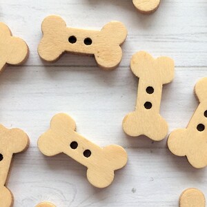 May include: A collection of wooden bone-shaped buttons with two holes each. The buttons are a light brown color and are arranged on a white surface.