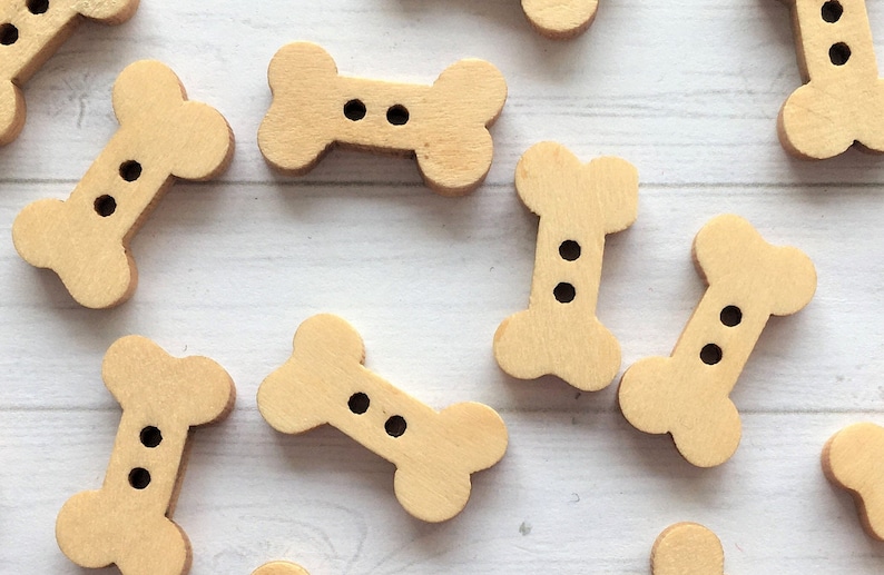May include: A collection of wooden bone-shaped buttons with two holes each. The buttons are a light brown color and are arranged on a white surface.