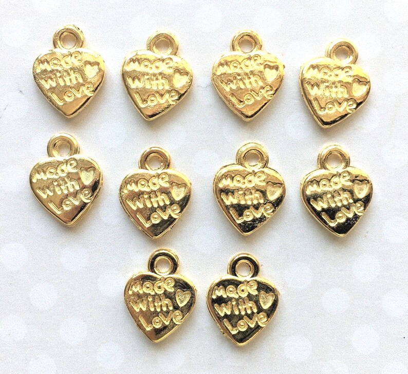 May include: A collection of ten gold-colored heart-shaped charms. Each charm has a loop at the top for attachment and the words "Made With Love" embossed on the front. The charms are suitable for jewelry making or craft projects.