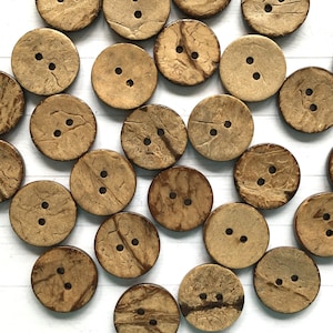 May include: A collection of round, natural coconut shell buttons in various shades of brown. The buttons have two holes each and are arranged in a random pattern.