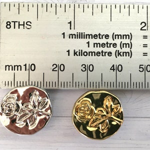 May include: Two round metal buttons with a raised design of a rose and leaves. One button is silver and the other is gold.
