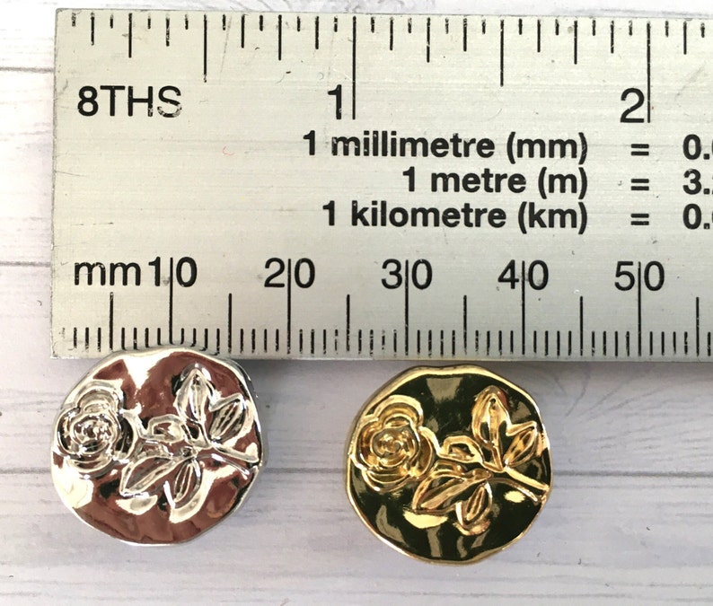 May include: Two round metal buttons with a raised design of a rose and leaves. One button is silver and the other is gold.