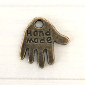 May include: A bronze-colored charm in the shape of a hand, with the words "Hand Made" inscribed on the palm. The charm has a small hole at the top for stringing or attaching to other items. The charm is likely used for jewelry making or crafting.