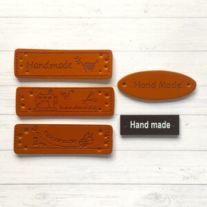 May include: Four brown leather labels with the text "Handmade" in different styles. One label is oval shaped and the other three are rectangular. One label has a sewing machine, scissors, and thread design. Another label has a bird and a flower design. The last label has a ball of yarn and a needle design. There is also a black rectangular label with the text "Hand made".