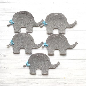 May include: Five grey felt elephant shapes with blue bows on their tails. The elephants are arranged in a semi-circle on a white wooden surface.