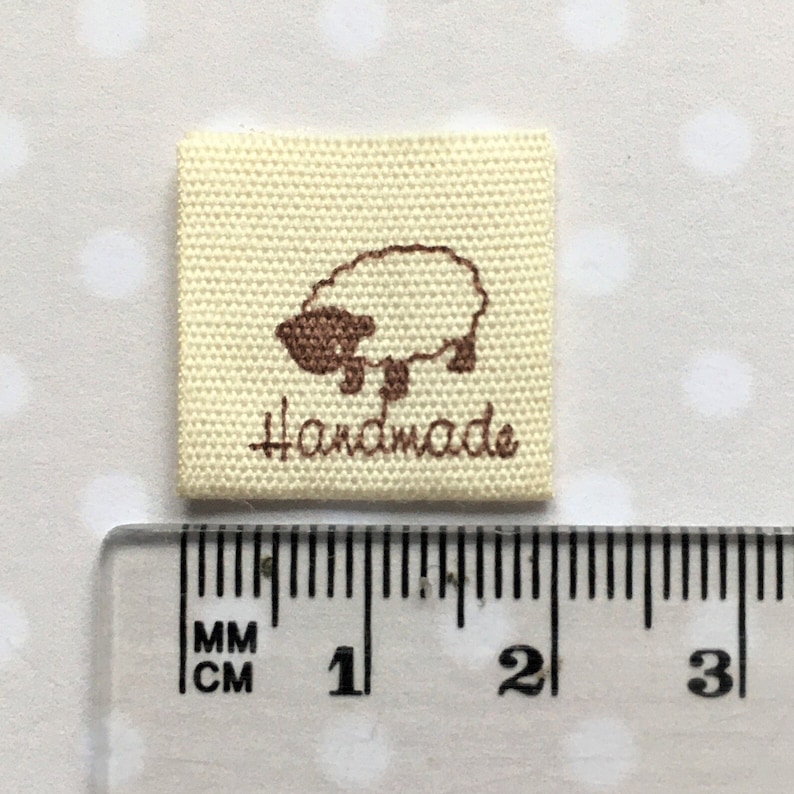 K&ouml;nnte beinhalten: Ein beigefarbenes Stofflabel mit einem braunen gestickten Schaf und dem Wort "Handmade". Das Label ist 2,5 cm quadratisch.