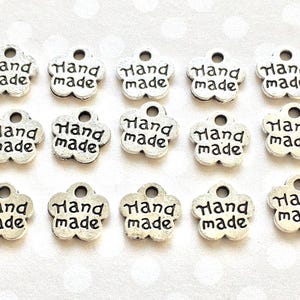 May include: A collection of silver-colored, flower-shaped charms. Each charm has the words "Hand made" in black lettering. The charms have a small hole at the top for stringing or attaching to other items. These charms are ideal for jewelry making or craft projects.