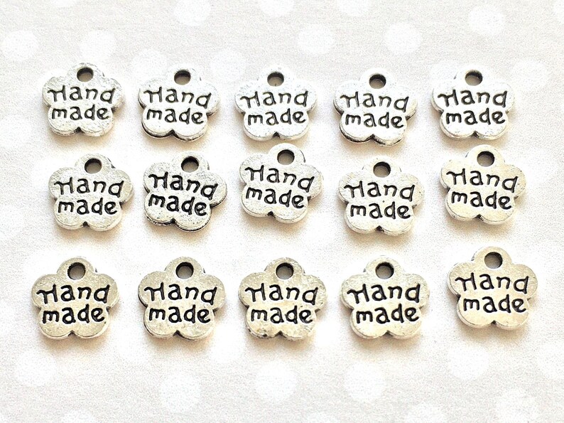 May include: A collection of silver-colored, flower-shaped charms. Each charm has the words "Hand made" in black lettering. The charms have a small hole at the top for stringing or attaching to other items. These charms are ideal for jewelry making or craft projects.