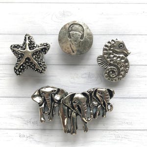 May include: Five silver-toned metal buttons featuring various designs. The buttons include a starfish, a seahorse, a portrait of a woman, and two elephants.