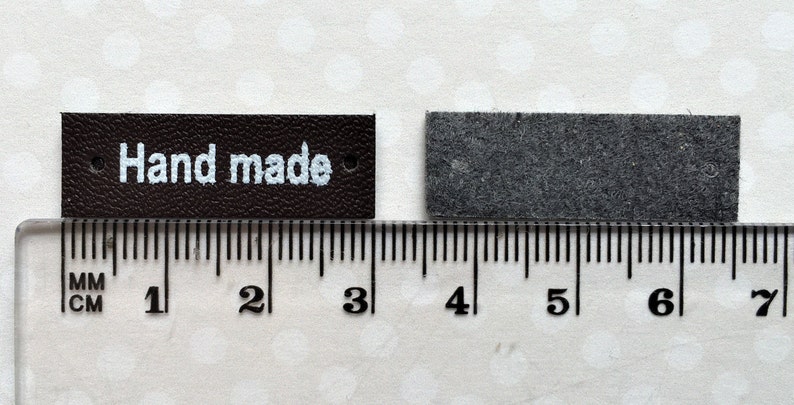 May include: Two brown leather labels with holes for stitching. The label on the left reads "Hand made" in white lettering. The labels are measured against a ruler with markings in centimeters and inches.