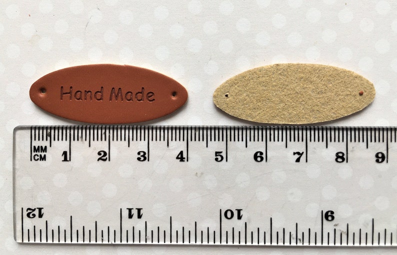 May include: Two oval-shaped leather tags, one brown with the text "Hand Made" and the other beige, both with two holes for stitching. The tags are measured against a ruler.