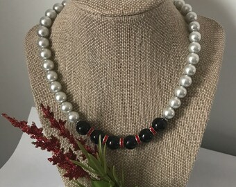May include: A white and black beaded necklace with red accents on a fabric-covered mannequin. The necklace is displayed against a white background.