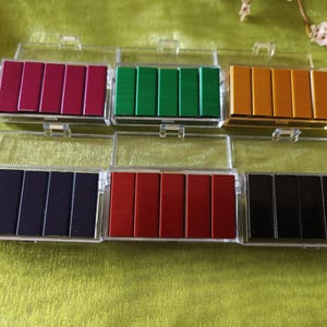 May include: A collection of colorful staple refills in clear plastic cases. The staples are arranged in rows of six and come in various colors, including red, green, gold, black, and navy blue. The cases have hinged lids.