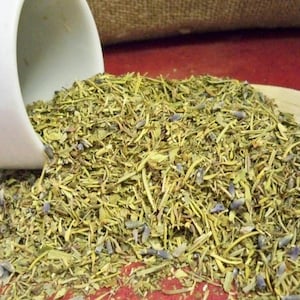 May include: A white ceramic cup pouring out a mixture of dried herbs and lavender flowers. The herbs are a mix of green and brown, and the lavender flowers are a light purple.