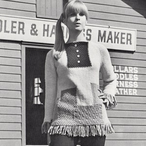 May include: A black and white photo of a woman wearing a knitted sweater with a patchwork design and fringe trim. The sweater has a high neckline and a button closure. The woman is standing in front of a building with a sign that reads "SADDLER & HARNESS MAKER".