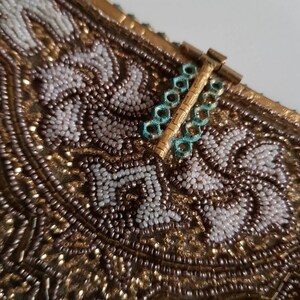 May include: Close-up of a vintage beaded handbag. The bag features intricate beadwork in gold, white, and brown tones, with a floral pattern. The clasp is gold with turquoise accents, adding a touch of colour to the design.