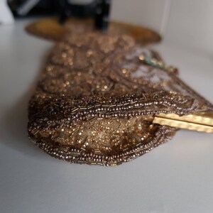 May include: A vintage gold beaded clutch bag with a rectangular shape. The bag features intricate beadwork and a gold clasp. The beads are in shades of gold and brown, creating a shimmering effect. The clutch is likely from the mid-20th century.