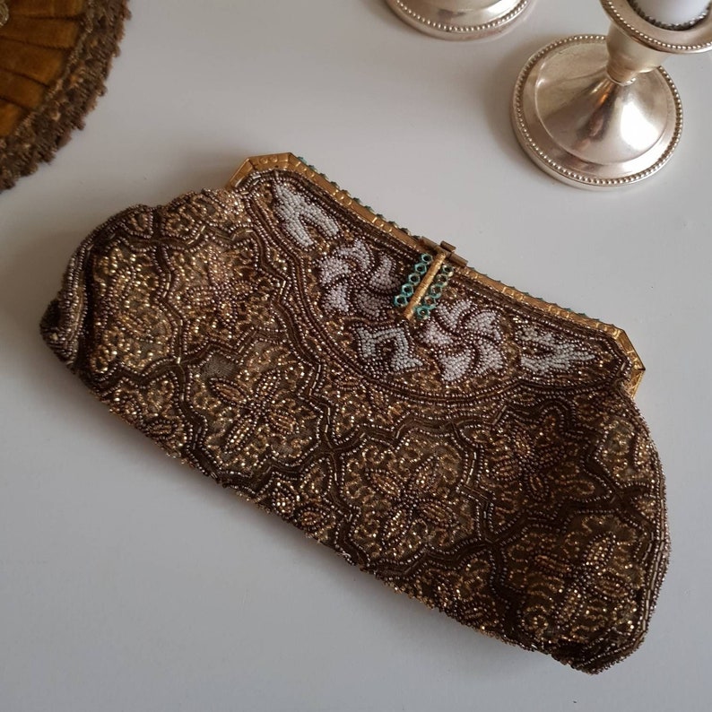 May include: A vintage beaded clutch bag in brown and gold, with detailed floral patterns. The top section has white beading and a gold frame with a turquoise clasp. A decorative item for special occasions.