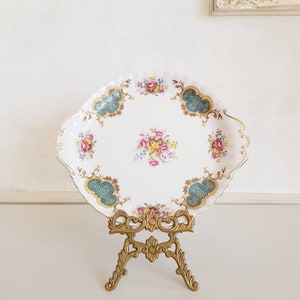 Op de afbeelding: Een witte ovale serveerschaal met een gouden rand en blauwe en gouden bloemmotieven. De schaal staat op een gouden metalen standaard.