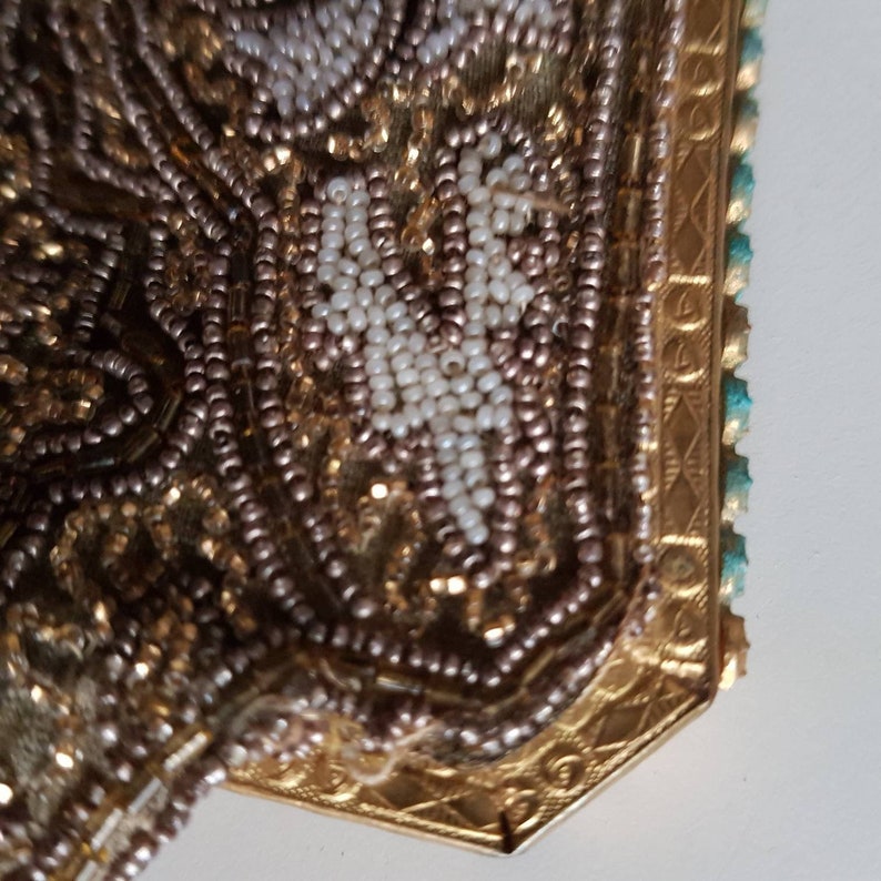 May include: Close-up of a vintage beaded purse. The purse features intricate beadwork in shades of gold, brown, and white. The frame is gold-toned with a decorative pattern, and the edge has turquoise accents. The purse is likely from the early to mid-20th century.