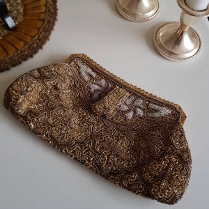 May include: A vintage beaded clutch bag in gold and brown tones, with detailed floral and geometric designs. The bag has a clasp closure and a gold frame. The top edge is decorated with white and gold beads. A classic accessory for special occasions.