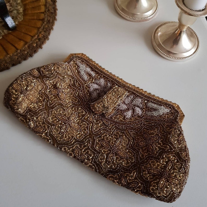 May include: A vintage beaded clutch bag in gold and brown tones, with detailed floral and geometric designs. The bag has a clasp closure and a gold frame. The top edge is decorated with white and gold beads. A classic accessory for special occasions.
