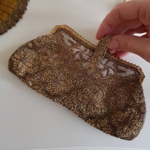 May include: A vintage beaded clutch bag in gold and brown tones. The bag is covered in detailed beadwork with a floral design and a small handle. The edges are trimmed with gold-coloured metal. The bag is held by a hand.
