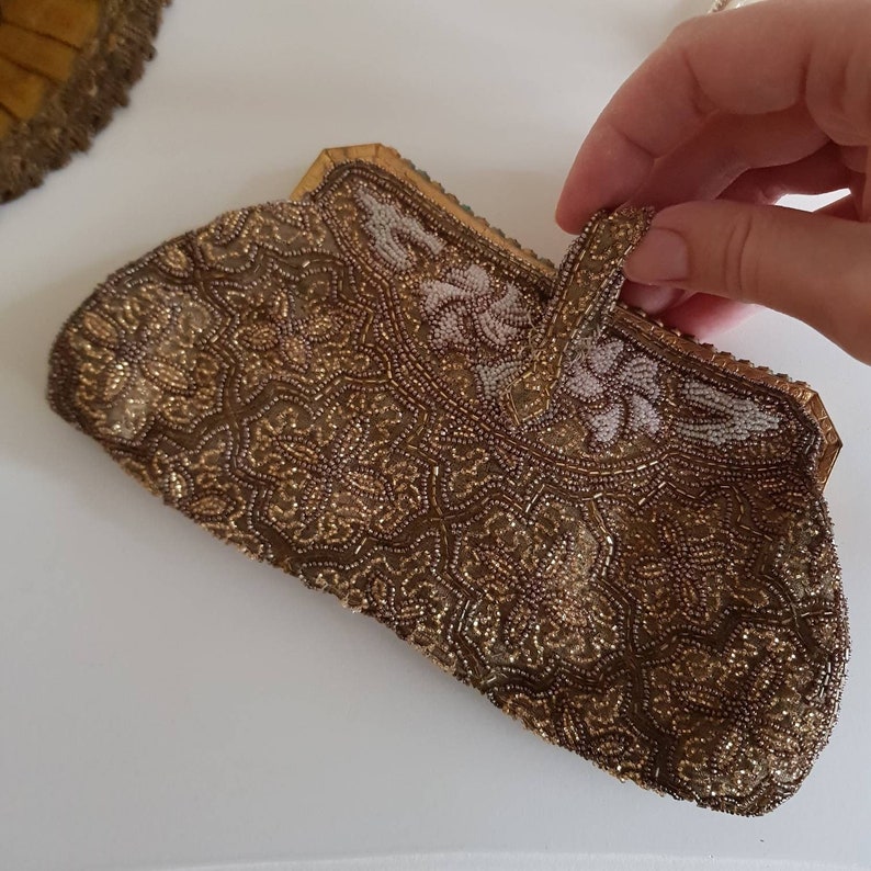 May include: A vintage beaded clutch bag in gold and brown tones. The bag is covered in detailed beadwork with a floral design and a small handle. The edges are trimmed with gold-coloured metal. The bag is held by a hand.