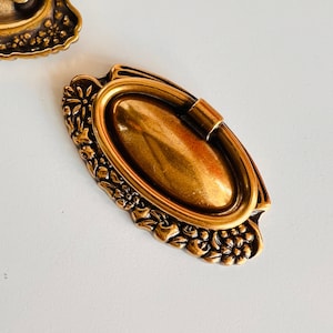 Op de afbeelding: Een ovale kastknop in goudkleur met een decoratief bloemmotief. De knop heeft een verhoogd ovaal midden met een glad oppervlak.