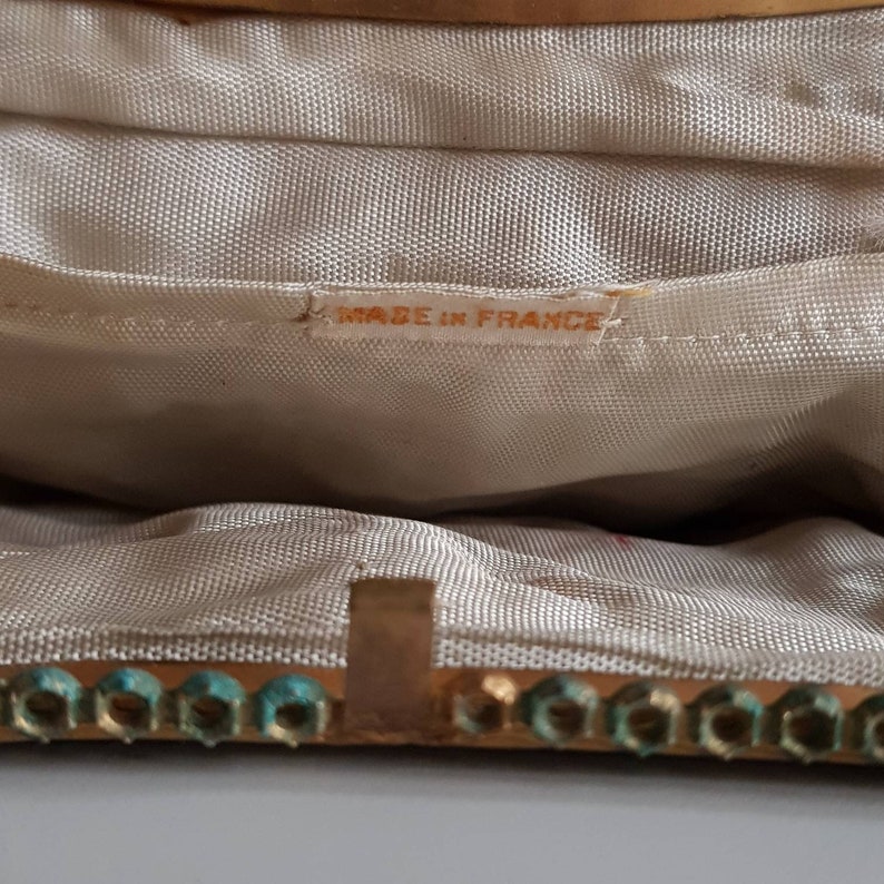 May include: Close-up of the interior of a vintage handbag. The lining is a light beige fabric. The gold-toned frame is embellished with turquoise-coloured accents. The label inside reads "MADE IN FRANCE".