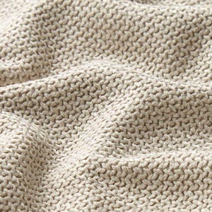 May include: Close-up of a beige knitted fabric with a textured, woven pattern. The fabric has a soft, undulating surface, with visible stitches creating a detailed, tactile appearance. The neutral color suggests a versatile textile suitable for various applications.