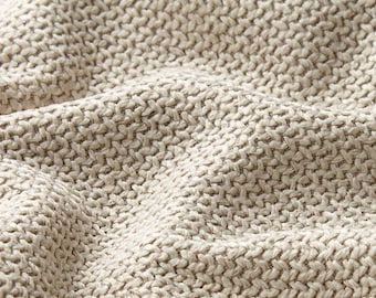 May include: Close-up of a beige knitted fabric with a textured, woven pattern. The fabric has a soft, undulating surface, with visible stitches creating a detailed, tactile appearance. The neutral colour suggests a versatile textile suitable for various applications.