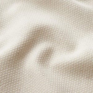 May include: Close-up of a textured, off-white fabric. The weave is visible, creating a subtle pattern of diagonal lines. The fabric has a soft, slightly rumpled appearance, suggesting a natural fibre textile.