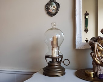 Può includere: Una lampada da tavolo in ottone vintage con un paralume in vetro trasparente e una singola candela all'interno. La lampada ha una maniglia a forma di chiave sul lato e si trova su un tavolo bianco.