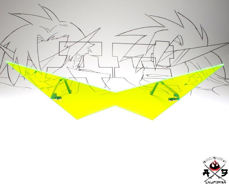 Puede incluir: Un par de gafas de sol triangulares amarillas con brazos verdes. Las gafas de sol est&aacute;n delante de un dibujo de dos personajes de anime.