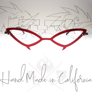 May include: Red cat-eye glasses with a sharp, angular design. The glasses are made of clear plastic and have a red frame. The glasses are reflected in a drawing of a cartoon character.
