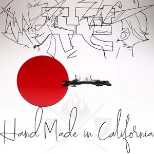 Puede incluir: Un círculo rojo con un clip negro unido a él. El clip es de metal y tiene un acabado negro. El fondo es blanco con un dibujo en blanco y negro de dos personas. El texto "Hand Made in California" está escrito en negro en la parte inferior de la imagen.