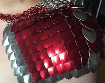 May include: A shoulder armour piece constructed from overlapping metallic scales in deep red and silver. The scales are connected by small metal rings, with a prominent red chainmail collar section encircling the neck area of the garment.
