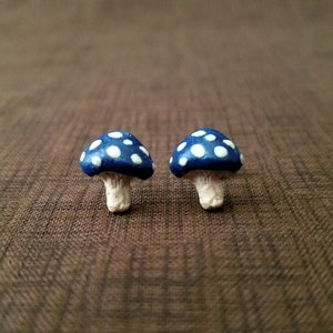 May include: Two small blue and white mushroom-shaped earrings. The mushrooms have white polka dots on a dark blue background.